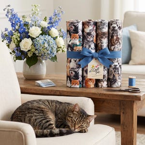 Könnte beinhalten: Ein Bündel Stoffrollen mit wiederkehrendem Katzenmuster, zusammengebunden mit einem blauen Band und einem Etikett mit der Aufschrift Quilt Shop Ruth Baudisch. Der Stoff zeigt verschiedene Katzenrassen in Braun-, Grau-, Weiß- und Orangetönen.