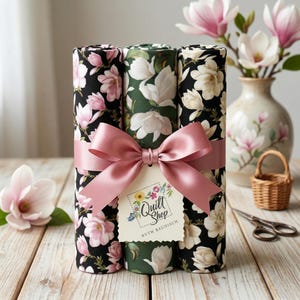 Pode incluir: Três rolos de tecido com estampas florais em preto, verde e branco, amarrados com uma fita rosa e uma etiqueta "Quilt Shop Ruth Baudisch". Um vaso de flores de magnólia e uma pequena cesta estão ao fundo. O tecido está sobre uma superfície de madeira.