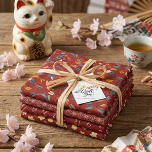 Peut inclure: Un ensemble de carrés de tissu rouge attachés avec du raphia, avec des motifs de libellules et géométriques. Une statue de chat Maneki-neko en céramique, des fleurs de cerisier et une tasse de thé sont également présents. L'étiquette indique "Quilt Shop, Ruth Baudisch".