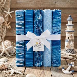 Può includere: Un fascio di sei rotoli di tessuto blu con motivi a tema oceano, legati con un nastro bianco e un'etichetta con la scritta "Quilt Shop". I tessuti presentano disegni di onde e acqua in varie tonalità di blu. Il fascio è esposto su una superficie di legno con decorazioni nautiche.