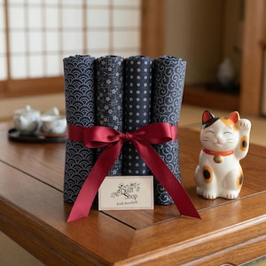 Può includere: Un fascio di quattro pezzi di tessuto blu navy arrotolati con vari motivi, legati con un nastro bordeaux. A destra c'è una statuina di gatto Maneki-neko in ceramica. Una piccola carta recita "Quilt Shop Ruth Baudisch."