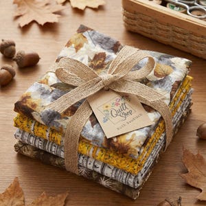 Könnte beinhalten: Ein Stapel Stoffquadrate mit Herbstmotiven, gebunden mit Schnur und einem Etikett vom Quilt Shop. Die Stoffe zeigen Blattmuster in Braun-, Grau- und Gelbtönen. Eicheln und getrocknete Blätter sind um das Stoffbündel herum verstreut.
