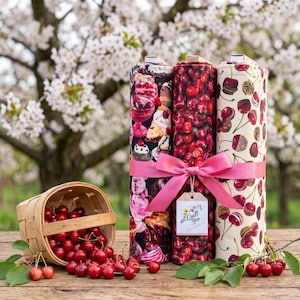 Peut inclure: Trois rouleaux de tissu attachés par un ruban rose avec une étiquette indiquant Quilt Shop Ruth Baurisch. Le rouleau de gauche présente des desserts, celui du centre un motif de cerises dense, et celui de droite des cerises sur fond crème avec un panier.