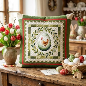 Op de afbeelding: Vierkant decoratief kussen met een Pasen thema. Het kussen heeft een centraal ontwerp van een ei met een kip erin, omringd door een groene krans. Het kussen heeft een groene rand met rode accenten en een crèmekleurige achterkant. Een vaas met tulpen en paaseieren staan in de buurt.