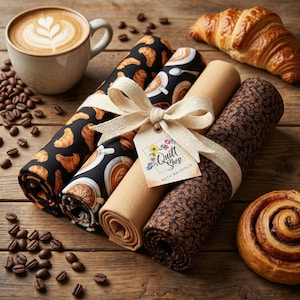 Könnte beinhalten: Ein Stillleben mit gerollten Stoffen mit Kaffee- und Gebäckmustern, gebunden mit einem Band und einem Etikett "Quilt Shop Ruth Baudisch". Eine Tasse Kaffee, Kaffeebohnen, ein Croissant und eine Zimtschnecke sind ebenfalls zu sehen.