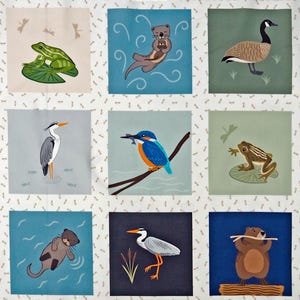 Op de afbeelding: Een patchwork quilt met geborduurde vierkanten met verschillende water- en bosdieren. De quilt toont een kikker op een waterlelie, een bever, een otter, een gans, een eend, een reiger, een ijsvogel en meer. Het kleurenpalet omvat blauw, groen en bruin.