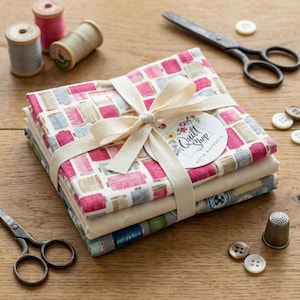Può includere: Un fascio di quadrati di tessuto, legato con un nastro color crema. Il tessuto presenta un motivo ripetuto di bobine rosa, grigie e beige. Un'etichetta con il testo "Quilt Shop" è attaccata. Accessori per cucire, tra cui forbici, bottoni e rocchetti di filo, sono sparsi.