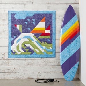 Puede incluir: Un edredón de pared cuadrado con un diseño geométrico de velero y olas en morado, verde y naranja, junto a una tabla de surf con rayas diagonales en morado, azul, amarillo, naranja y rojo, y una correa de tabla de surf negra enrollada.