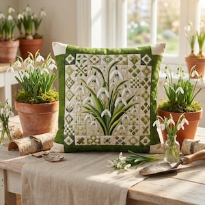 Puede incluir: Un cojín cuadrado decorativo con un diseño de patchwork de flores de campanillas de invierno. El cojín tiene un borde verde y una parte trasera crema. Las campanillas de invierno en macetas de terracota y pequeños jarrones de cristal están dispuestas alrededor.