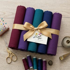 Op de afbeelding: Een bundel gekleurde stoffen rollen in tinten bordeauxrood, paars, teal en marineblauw, vastgebonden met een gouden lint en strik. Een label met de tekst "Quilt Shop Ruth Baudisch" is bevestigd. Naaigerei, waaronder schaar, spelden en garenspoelen, zijn ook aanwezig.