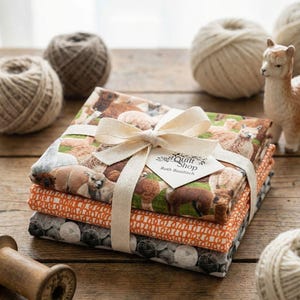 Peut inclure: Un ensemble de tissus empilés, liés par un ruban crème. Le tissu supérieur présente un motif d'alpagas. Les autres tissus ont des motifs orange et gris. Une étiquette indique "Quilt Shop Ruth Baudisch". Des boules de fil et un alpaga en céramique sont en arrière-plan.