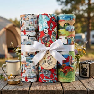 Può includere: Tre rotoli di tessuto legati con un nastro bianco e un'etichetta. I rotoli presentano motivi a tema campeggio con roulotte vintage, fotocamere e paesaggi. Una fotocamera vintage, una bussola e una tazza sono su un tavolo di legno.