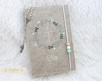 May include: A beige felt cover featuring an embroidered floral wreath surrounding a central cross. The name Ella is embroidered in white below the wreath. A white elastic band with three wooden beads in tan, white, and mint green secures the cover.