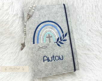 May include: A light gray felt book cover featuring an embroidered blue rainbow, a cross, a fish symbol, and a leafy branch. The name Anton is embroidered in blue thread at the bottom. A dark blue elastic band secures the cover, and a silver rosary is attached.