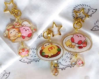 May include: Four keychains featuring cartoon characters from the video game Kirby. Each keychain has a different design, including Kirby eating a meal, Kirby sleeping, and Kirby with a friend. The keychains are made of clear acrylic and have gold metal rings.