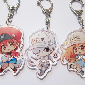 Könnte beinhalten: Drei Anime-Charakter-Schlüsselanhänger mit klaren Acryl-Rückseiten. Die Charaktere tragen rote, weiße und rosa Hüte mit dem Text "白血球", "血小板" und "sample" darauf. Die Charaktere halten verschiedene Gegenstände, darunter ein Buch, eine Spritze und einen Verband.