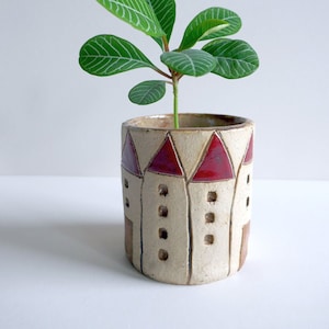 Könnte beinhalten: Ein kleiner, weißer Keramiktopf mit einem roten und braunen Design, das an eine Reihe von Häusern erinnert. Eine grüne Pflanze mit dünnen, gemusterten Blättern wächst im Topf.