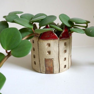 Könnte beinhalten: Kleiner, beigefarbener Keramik-Pflanztopf in Form eines Schlosses mit rotem Dach. Der Pflanztopf hat eine braune Tür und quadratische Fenster. Grüne, belaubte Pflanzenstiele und Blätter ragen aus der Oberseite des Pflanztopfes heraus. Der Pflanztopf steht auf einer weißen Oberfläche.