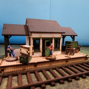 May include: A wooden model train station with a brown roof, white walls, and green trim. The station has a platform with a wooden floor and a railing. There are three miniature figures on the platform, one of which is sweeping the floor. The station is set on a track with brown wooden ties.
