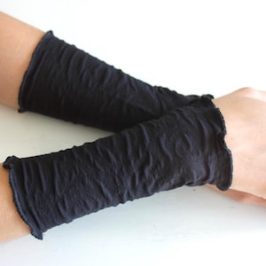 K&ouml;nnte beinhalten: Schwarze Armstulpen aus strukturiertem, gekr&auml;useltem Stoff. Die Armstulpen haben einen ger&uuml;schten Rand. Das Bild zeigt die Armstulpen an den Armen einer Person.