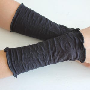 K&ouml;nnte beinhalten: Ein Paar dunkelgraue Armstulpen mit strukturiertem, zerknittertem Aussehen. Die Armstulpen haben einen ger&uuml;schten Rand oben und unten. Das Bild zeigt die Armstulpen an den Armen einer Person, die auf einer wei&szlig;en Oberfl&auml;che ruhen.
