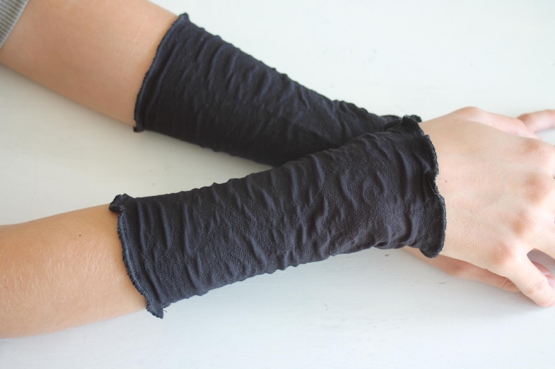 K&ouml;nnte beinhalten: Ein Paar dunkelgraue Armstulpen mit strukturiertem, zerknittertem Aussehen. Die Armstulpen haben einen ger&uuml;schten Rand oben und unten. Das Bild zeigt die Armstulpen an den Armen einer Person, die auf einer wei&szlig;en Oberfl&auml;che ruhen.