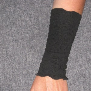 K&ouml;nnte beinhalten: Eine schwarze Handgelenkmanschette aus strukturiertem Stoff. Die Manschette wird am Handgelenk und Unterarm getragen, mit einem ger&uuml;schten Rand oben. Der Hintergrund ist einfarbig grau.