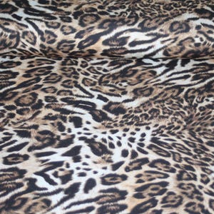 May include: A leopard print fabric. The material displays a repeating pattern of brown and black spots and stripes against a cream backdrop. The fabric is rolled, revealing the repeating design.