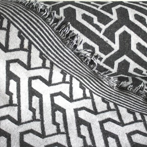 May include: A gray and white geometric patterned throw blanket. The blanket features a repeating angular design in shades of gray and white. A striped border with fringe adds a decorative touch to the blanket.