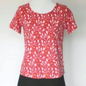Könnte beinhalten: Rotes kurzärmeliges Oberteil mit weißem Blumenmuster und Rundhalsausschnitt. Das Oberteil wird mit einem schwarzen Rock kombiniert. Das florale Design umfasst weiße Blätter und orangefarbene Akzente auf rotem Hintergrund. Kleidung auf einer Schaufensterpuppe.
