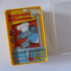Könnte beinhalten: Eine gelbe Vintage-Quartett-Kartenspielbox von Benjamin Blümchen. Die Box zeigt eine Illustration eines Elefanten mit Kochmütze und Schürze, der in einer Küche kocht. Der Text "Benjamin Blümchen" ist auf der Oberseite der Box aufgedruckt.
