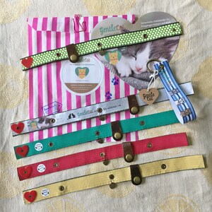 May include: Five colorful cat collars with a heart-shaped charm and a bell. The collars are made of fabric and have a safety buckle. The collars are displayed on a striped pink and white background. The text "Gentletat.de" is visible on one of the collars.