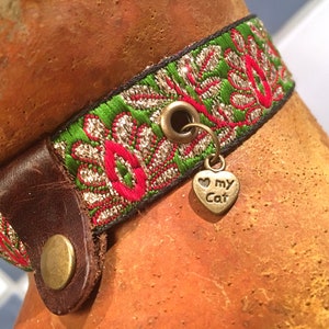 May include: A green, red, and gold patterned cat collar with a brown leather buckle and a heart-shaped charm that says "I love my cat".