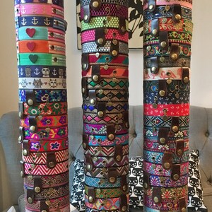 May include: Three stacks of colorful bracelets with different patterns and designs. Each stack is held together by a brown leather strap with metal buttons. The bracelets are made of fabric and have a variety of charms and embellishments.