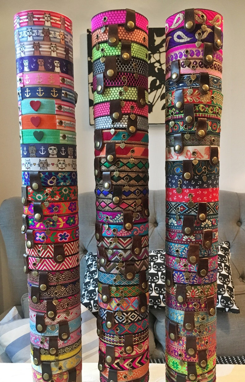 May include: Three stacks of colorful bracelets with different patterns and designs. Each stack is held together by a brown leather strap with metal buttons. The bracelets are made of fabric and have a variety of charms and embellishments.