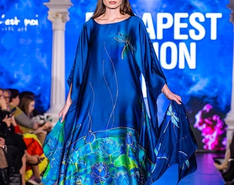 May include: A blue silk dress with a dragonfly and lotus flower design. The dress has long sleeves and a flowing skirt. The model is wearing the dress on a runway with a blue and white background.