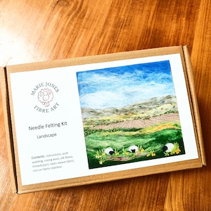 May include: A needle felting kit for creating a landscape scene. The kit includes instructions, roving wool, silk fibers, threads, open weave fabric, and iron-on fabric stabilizer. The image on the box shows a green field with sheep and a mountain range in the background.