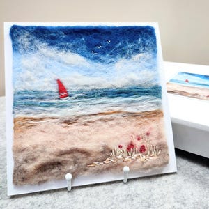May include: A needle felted artwork depicting a seascape with a red sailboat on the water, a sandy beach, and a small cluster of red flowers in the foreground.