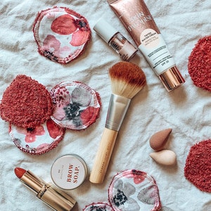 May include: An assortment of makeup products and accessories, including a tube of Skin Paradise tinted water-cream, nail polish, makeup brushes, reusable makeup remover pads with floral patterns, and a lipstick. The items are arranged on a white, textured surface.
