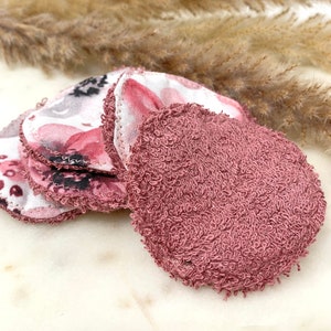 May include: A collection of reusable facial rounds. Some rounds feature a floral pattern in pink, white, and gray, while others are a solid mauve color. The rounds are made of soft, absorbent fabric, ideal for skincare routines.