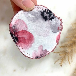 May include: A round, reusable cotton pad with a floral pattern in shades of burgundy, gray, and black. The edges are finished with a burgundy stitch. The pad is designed for skincare and makeup removal.