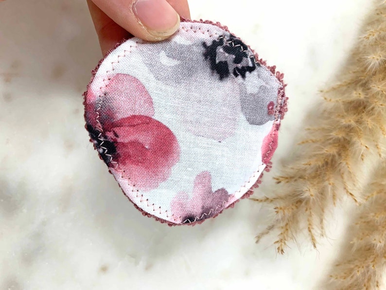 May include: A round, reusable cotton pad with a floral pattern in shades of burgundy, gray, and black. The edges are finished with a burgundy stitch. The pad is designed for skincare and makeup removal.