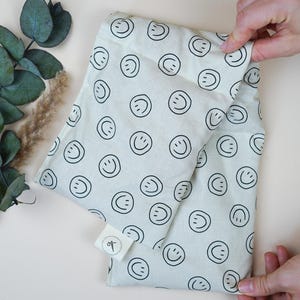 May include: A white fabric pouch with a black smiley face pattern. The pouch is folded in half and has a small white label with a black design on it.