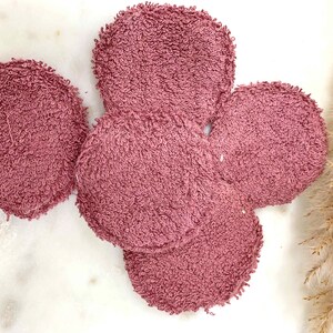 May include: A collection of five round, mauve-colored reusable facial cleansing pads. The pads are made of a soft, textured material, likely terry cloth, and are arranged on a white marble surface. The pads are a light, dusty rose color.