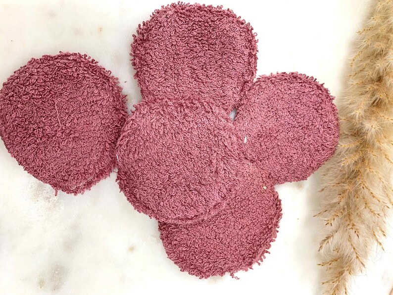 May include: A collection of five round, mauve-colored reusable facial cleansing pads. The pads are made of a soft, textured material, likely terry cloth, and are arranged on a white marble surface. The pads are a light, dusty rose color.