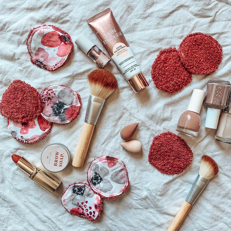 May include: An assortment of makeup products and accessories, including a tube of Skin Paradise tinted water-cream, nail polish, makeup brushes, reusable makeup remover pads with floral patterns, and a lipstick. The items are arranged on a white, textured surface.