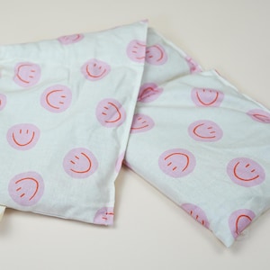 Könnte beinhalten: Zwei rechteckige Kissen aus weißem Stoff mit aufgedruckten pinkfarbenen Smiley-Gesichtern. Die Kissen sind aus einem weichen, leichten Stoff gefertigt und eignen sich perfekt, um jedem Raum eine Prise Verspieltheit zu verleihen.