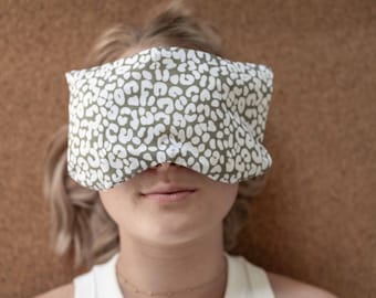 以下が含まれることがあります： 緑と白の斑点模様のアイピロー。白いタンクトップと金のネックレスを身につけた人の目をアイピローが覆っています。背景は無地の茶色です。