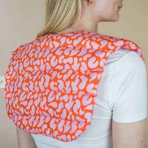 May include: A pink and orange patterned heat pack designed for shoulder and neck pain relief. The pack is made of soft fabric and has a quilted design.