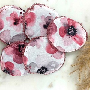 May include: A collection of six round, reusable facial pads. Each pad features a watercolor floral design in shades of pink, gray, and black on a white background. The edges are finished with a ruffled, mauve trim. These pads are designed for skincare.